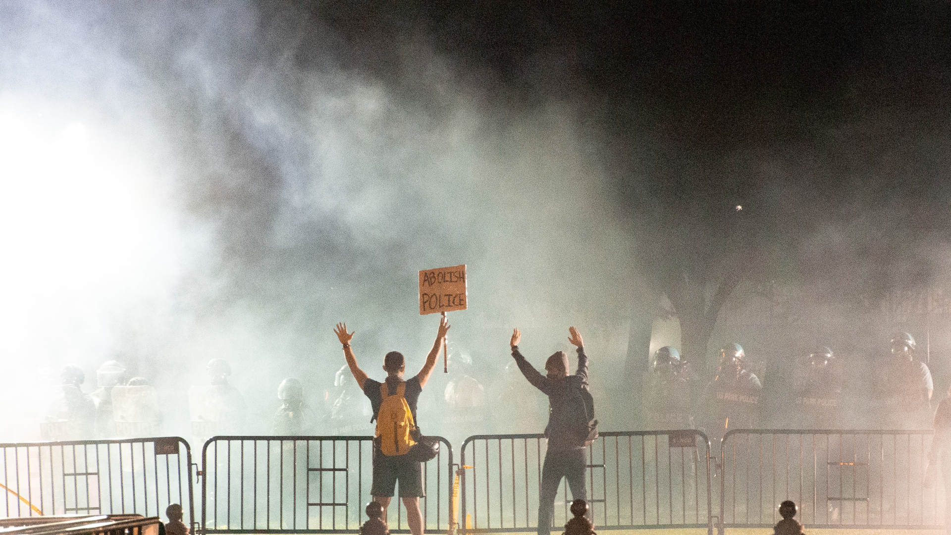 Two protesters hold up their hands in front of a line of police, with a cloud of tear gas wafting in front of them.