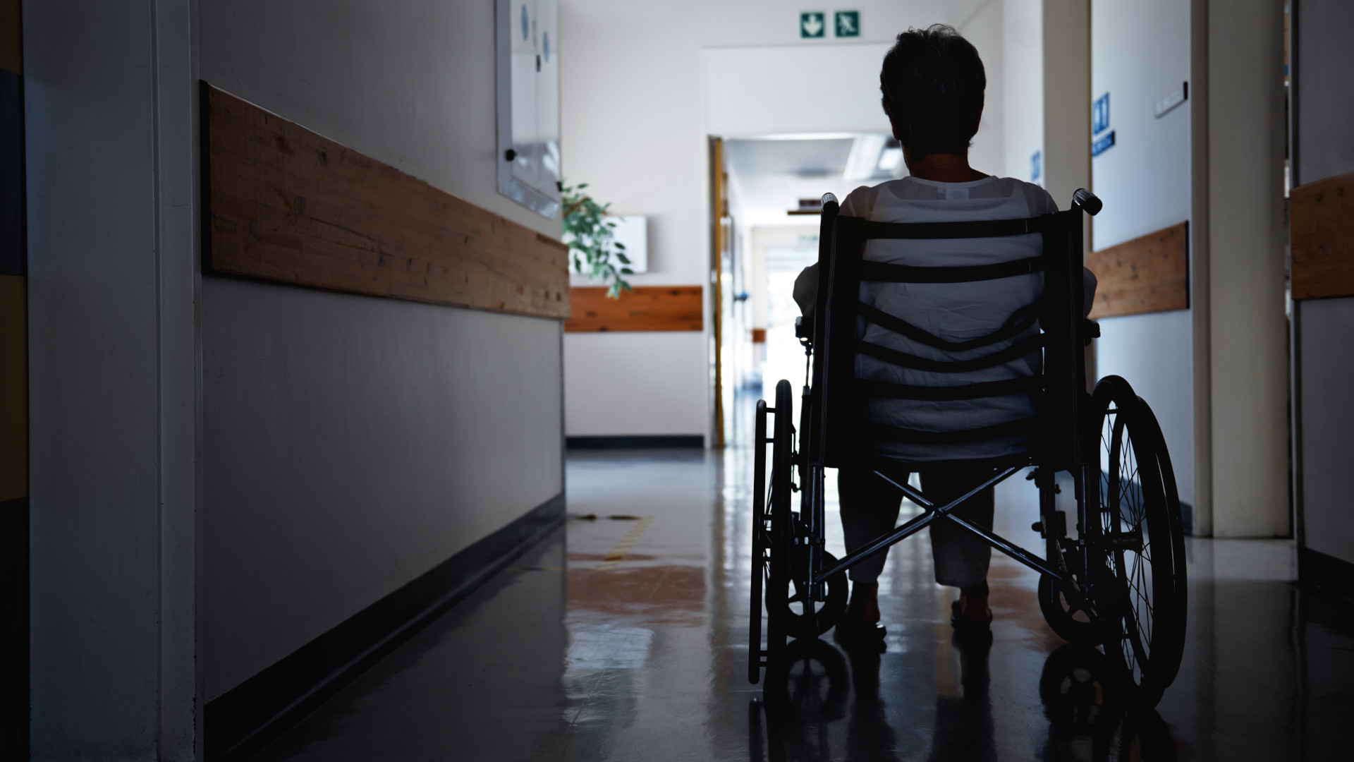 a woman, facing away from the camera, sits in a wheelchair in a dark hospital hallway