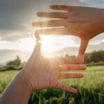 Close-Up of Female Hand Outstretched Touching Scenic View Of Mountain Range During Sunset.