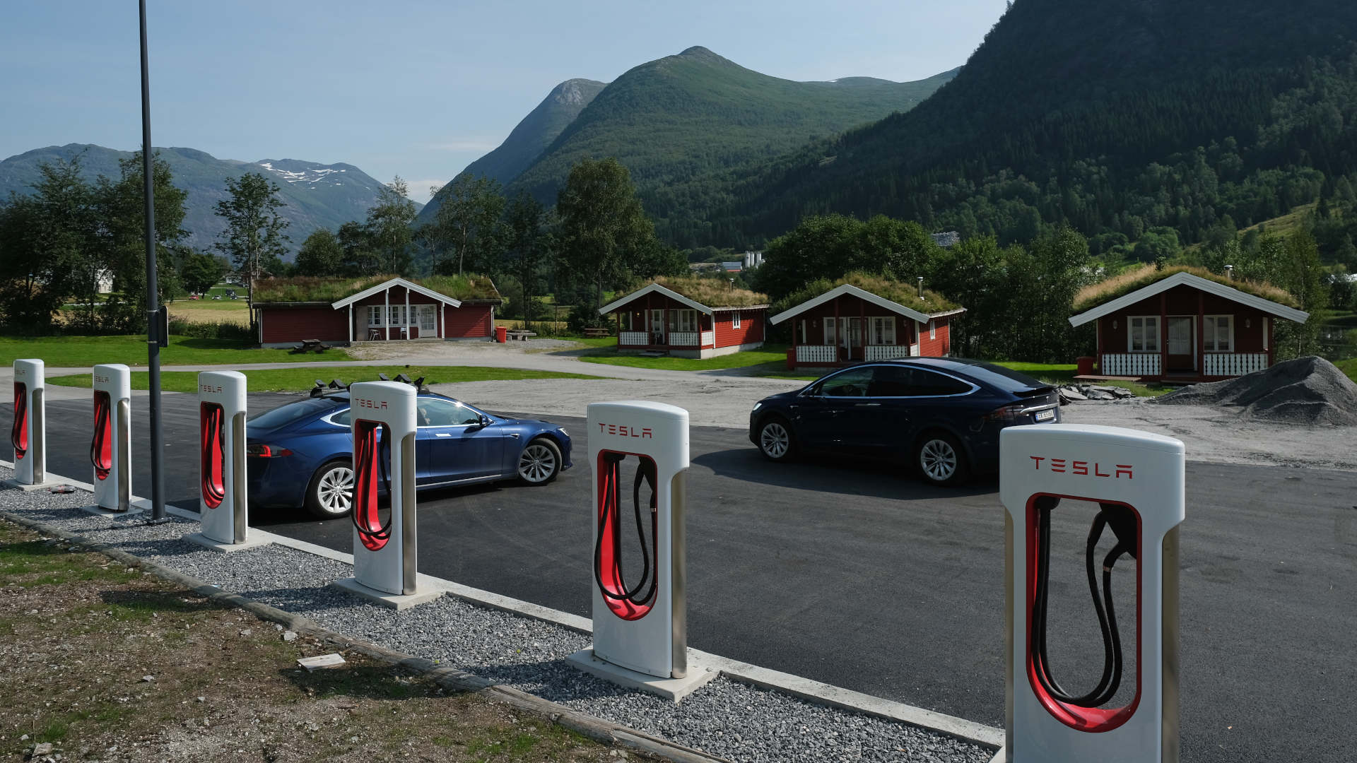 a parking lot with Tesla chargers sits next to several small red cabins with green mountains in the background.