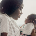A mother holds her newborn baby in her arms as she sits up in her hospital bed. She is Black nd wearing a hospital gown and is gazing down at her baby.