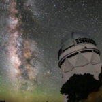A white structure with a domed top on a rocky outcrop below a star-filled night sky. The Milky Way stretches vertically in a luminous band. Faint green glow tints the curved horizon.