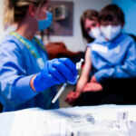 nurse in blue scrubs reaches for a syringe in the foreground while a mother and child sit in the background