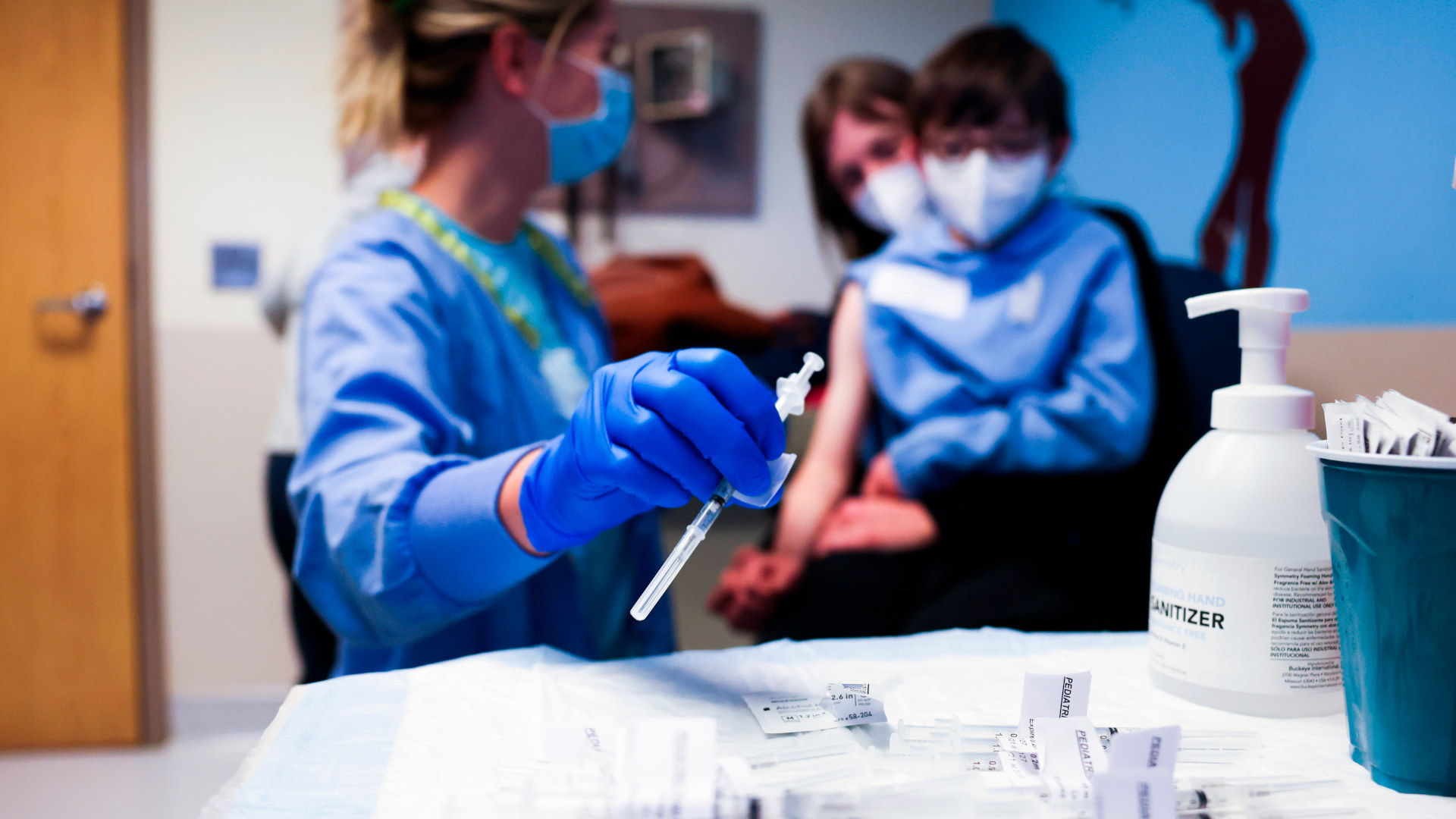 nurse in blue scrubs reaches for a syringe in the foreground while a mother and child sit in the background