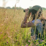 a woman looks through binoculars in a field of grass that nearly towers over her