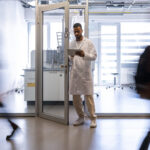 A chemical worker in a white coat stands between the doors of the faculty laboratory, students rush past him in the corridor. Blurred people while moving on the photo.