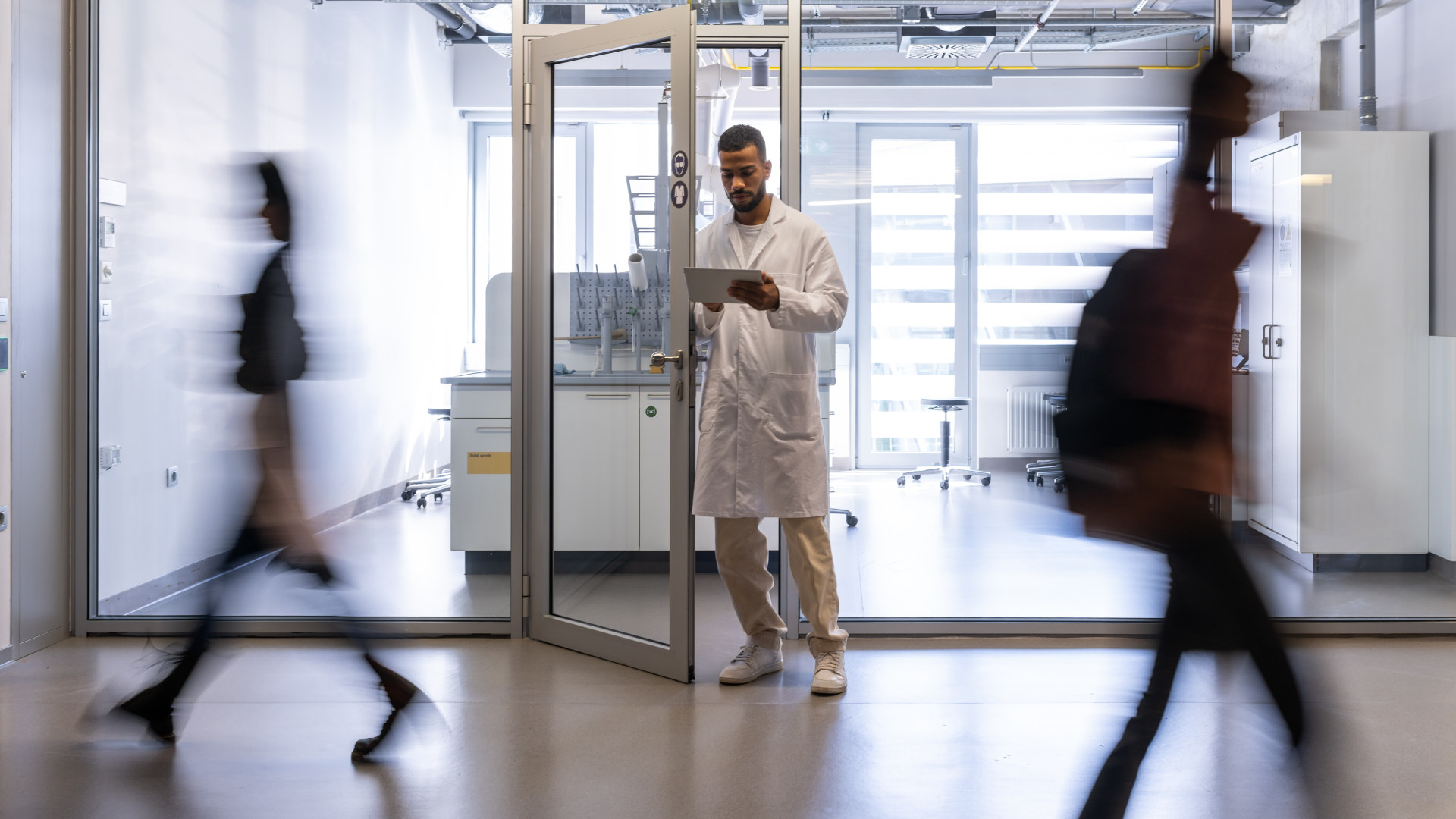 A chemical worker in a white coat stands between the doors of the faculty laboratory, students rush past him in the corridor. Blurred people while moving on the photo.
