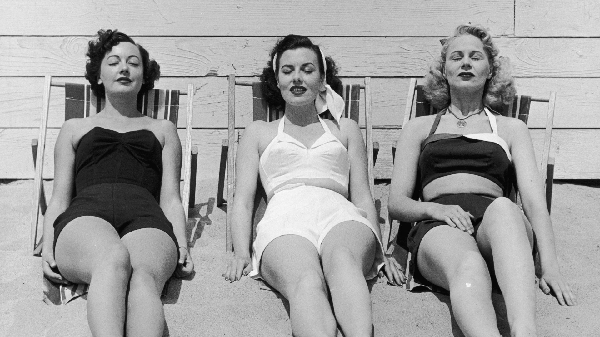 circa 1955: Three women in bathing suits sunbathe against a wall on a beach, early 1950s.