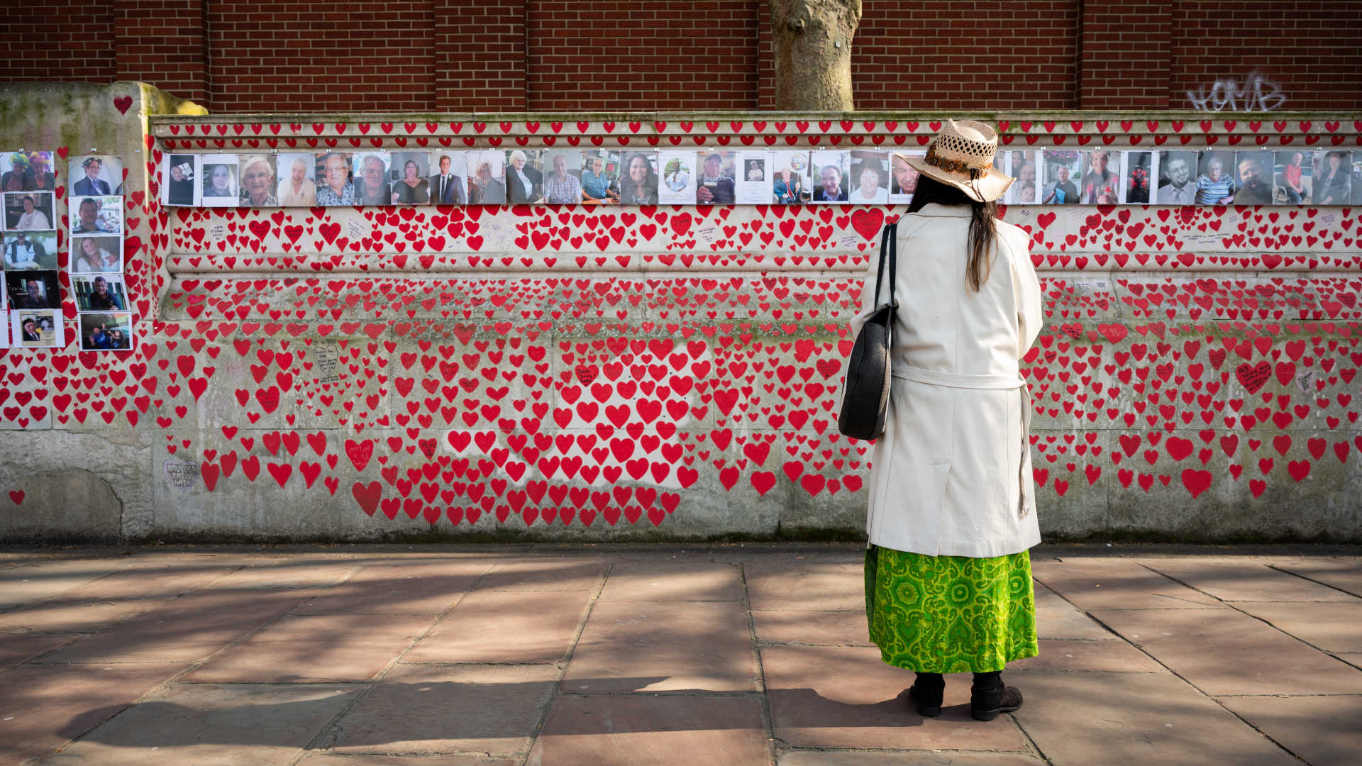 a woman stand in front of a wall decorated with red hearts and photos of people who died during the Covid-19 pandemic