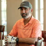 a man sits at a dining table. he wears an orange shirt and baseball cap.