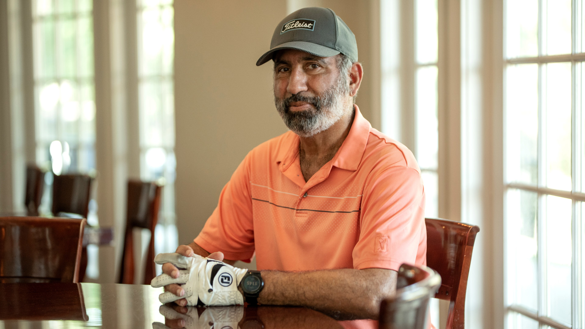 a man sits at a dining table. he wears an orange shirt and baseball cap.