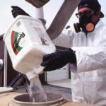 Farm worker wearing a respirator, a protective white suit, and black gloves pours Roundup into a tank.
