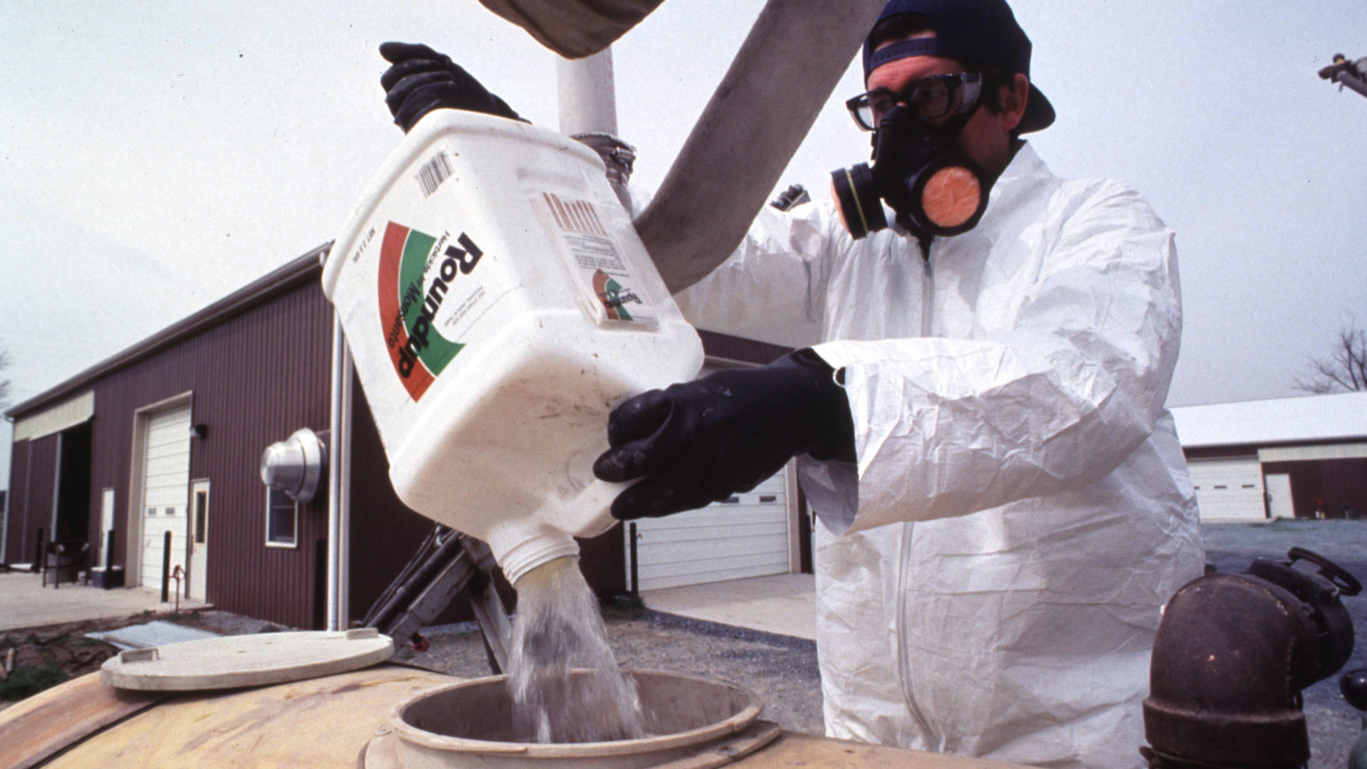 Farm worker wearing a respirator, a protective white suit, and black gloves pours Roundup into a tank.