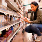 A woman reads the back of boxed pasta on the lower shelf in the grocery store aisle.