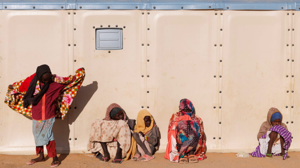Sudanese refugees sit and stand outside of a pale khaki structure