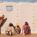 Sudanese refugees sit and stand outside of a pale khaki structure