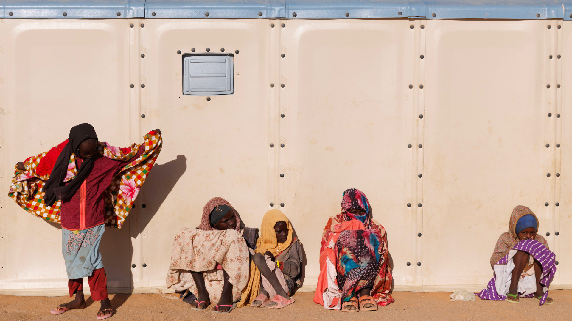 Sudanese refugees sit and stand outside of a pale khaki structure