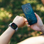 Over the shoulder view of active Asian sports woman wearing smartwatch and using fitness app on smartphone to monitor training progress after working out outdoors. Health and fitness training with technology. Wearable technology. Healthy living lifestyle