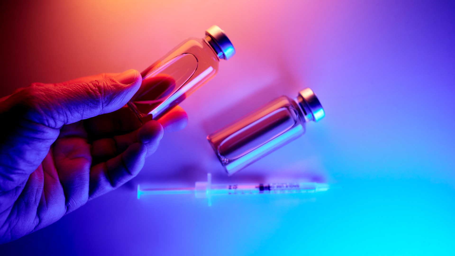 Hand holding a medication vial, two vials, and an injection syringe on a vibrant background.