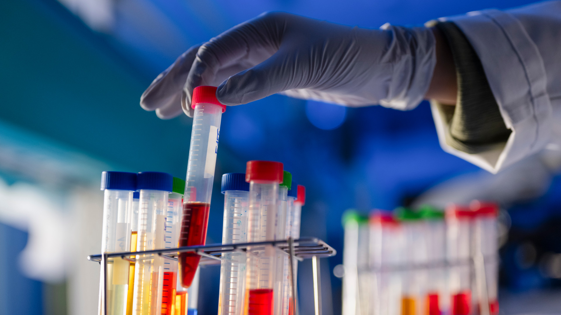 Test tubes filled with colorful samples in the lab. A glove handed grabs one from the rack.